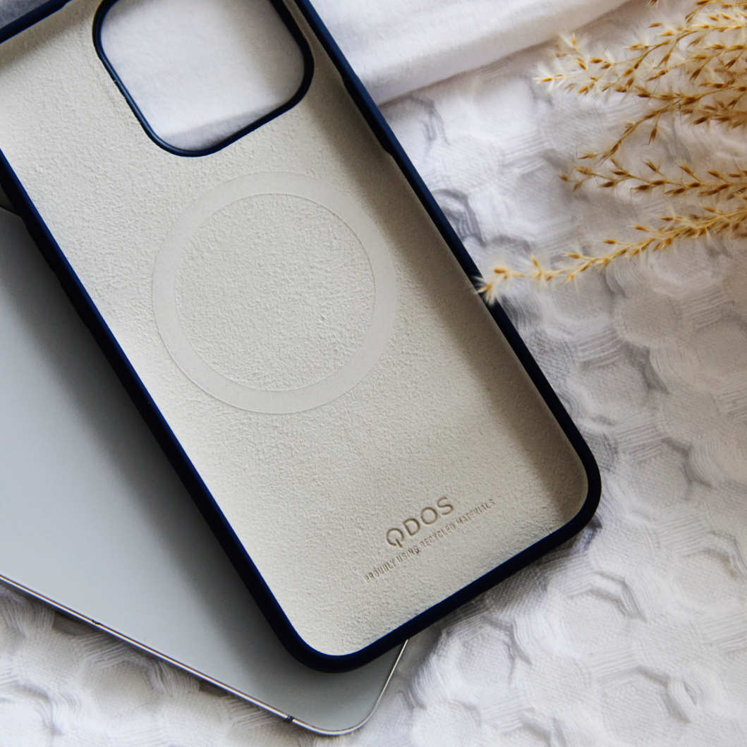 A close-up of the inside of a QDOS phone case, showing the soft inner lining and MagSafe compatibility ring. The case is resting on a textured white fabric next to dried wheat stems, creating a minimalistic, natural scene.