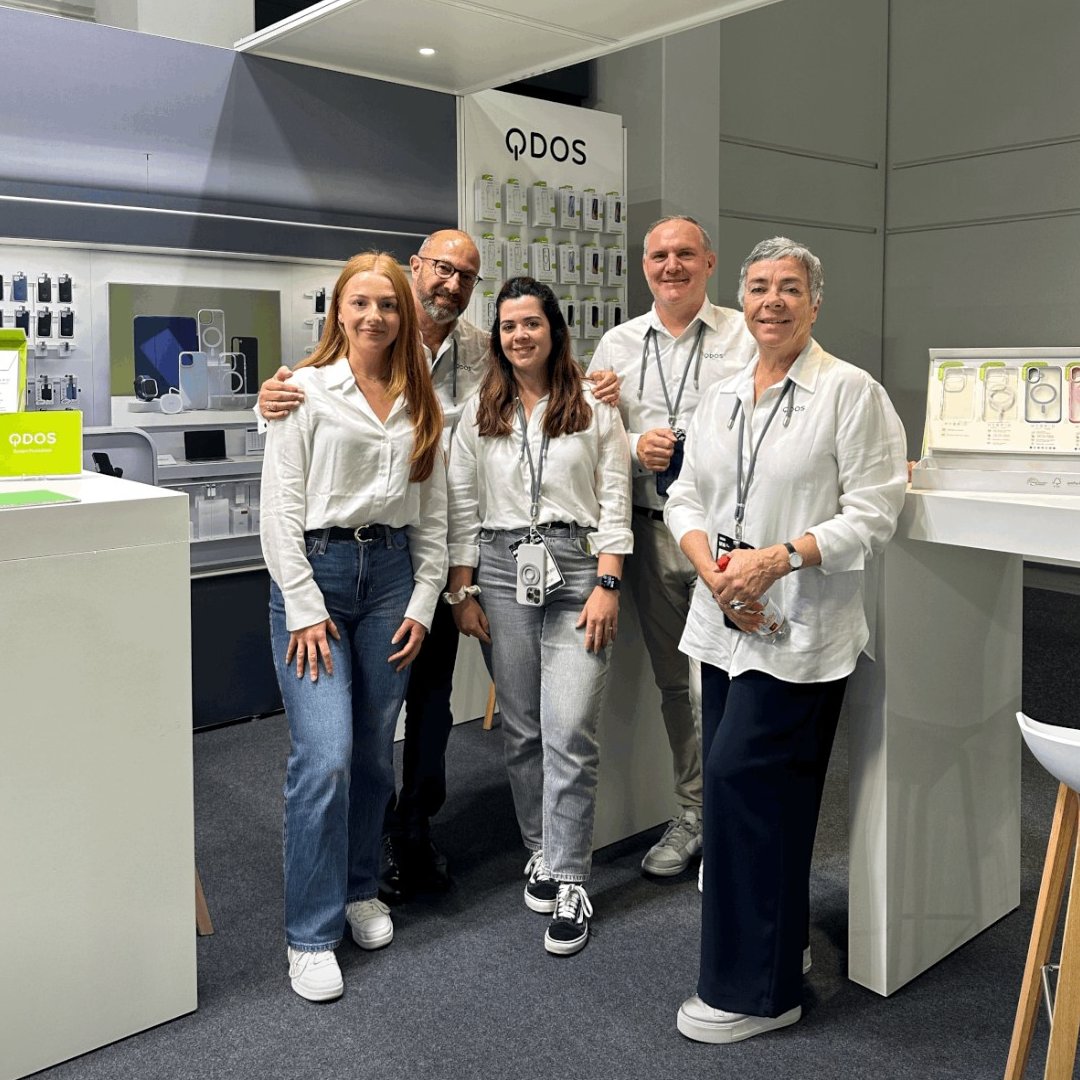 This image shows five team members from QDOS standing together in a trade show booth. They are dressed in coordinated white shirts and casual attire. The booth displays QDOS products such as phone cases and tech accessories. The group smiles warmly for the camera.