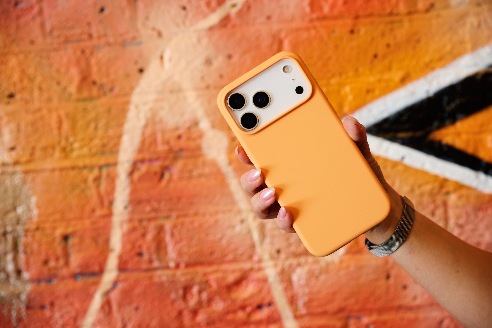 A hand holding an iPhone with a bright orange protective case, set against a blurred brick wall background.