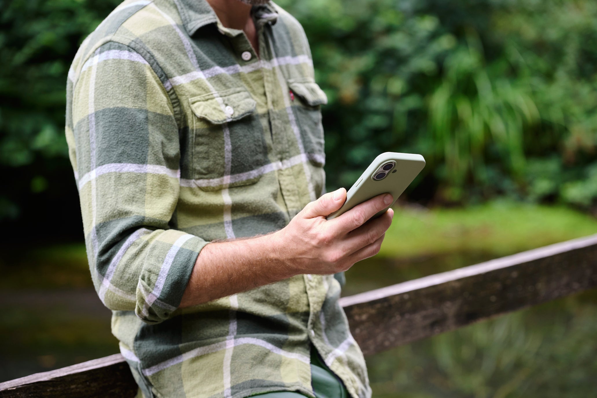 Person wearing a green plaid shirt holding a phone outdoors with blurred greenery in the background
